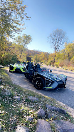 Two sporty three-wheeled open-top roadsters parked on a scenic country road amid fall foliage and clear blue sky