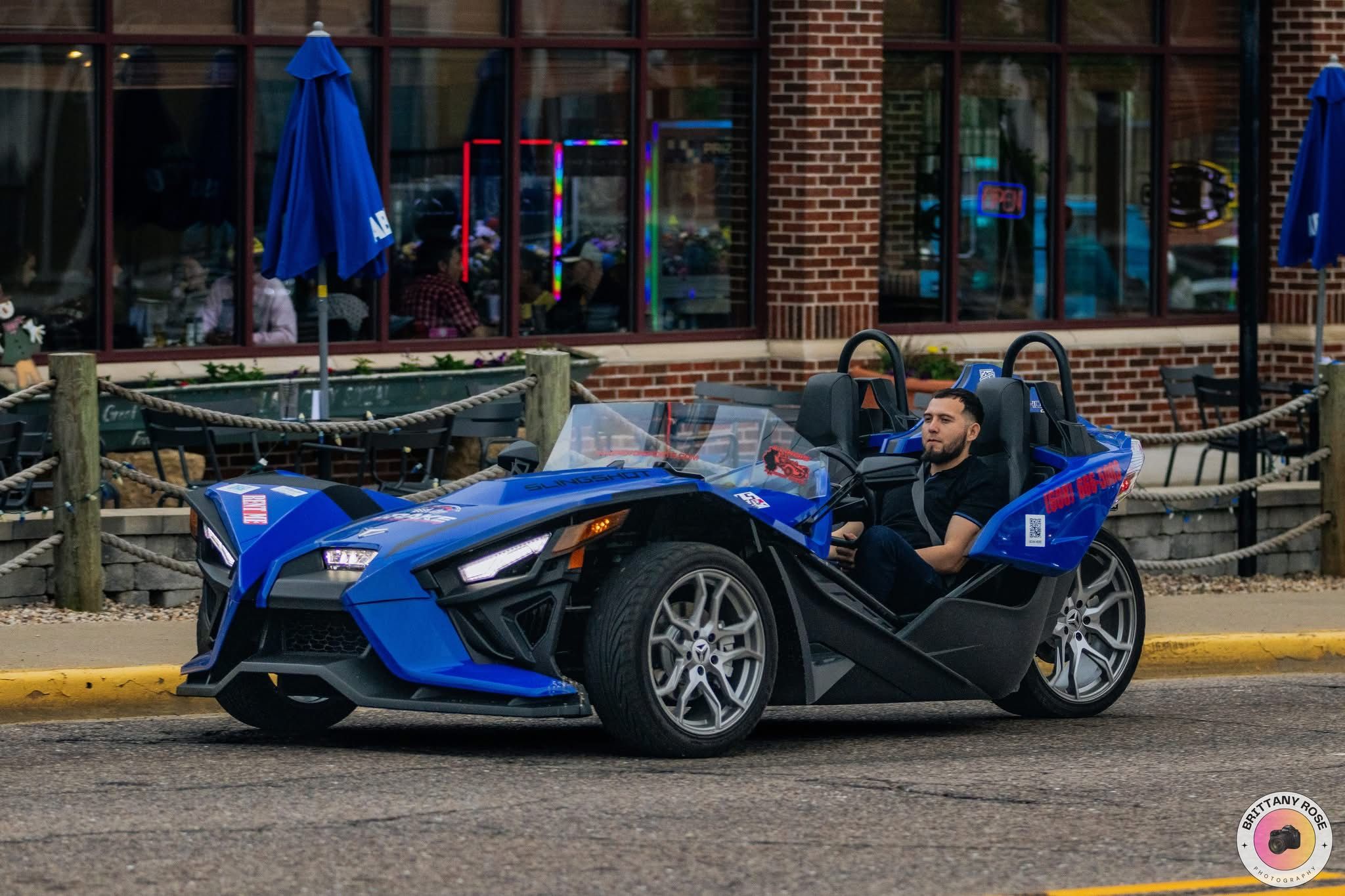 Eye-catching blue three-wheeled open-air roadster driven along a downtown street past a brick storefront with outdoor cafe seating and blue umbrellas.