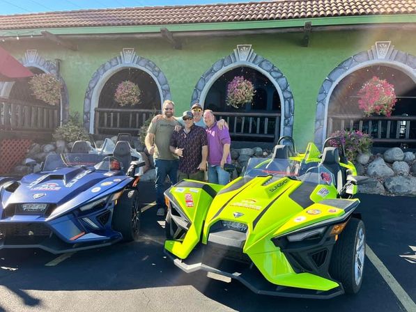 Four people posing between a neon green and deep blue Polaris Slingshot three-wheeled roadsters parked in a sunlit lot in front of a green stucco building with arched windows and hanging flower baskets.