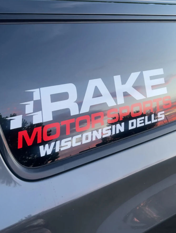 Bold white-and-red motorsports decal on a car rear window reading Wisconsin Dells, with a colorful sunset reflection in the glass
