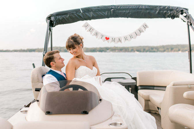 Newlywed couple on a pontoon boat with a "Just Married" banner, bride in a strapless white gown and groom in a blue vest smiling on a calm lake.