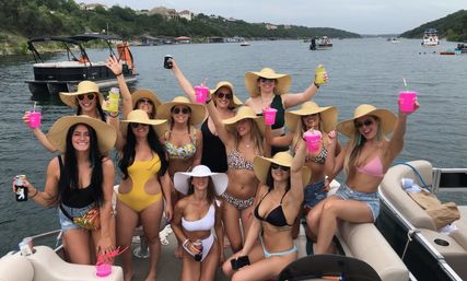 Group of women in swimsuits and wide-brim hats cheering with colorful cups on a pontoon boat at a sunny lake with shoreline homes and other boats