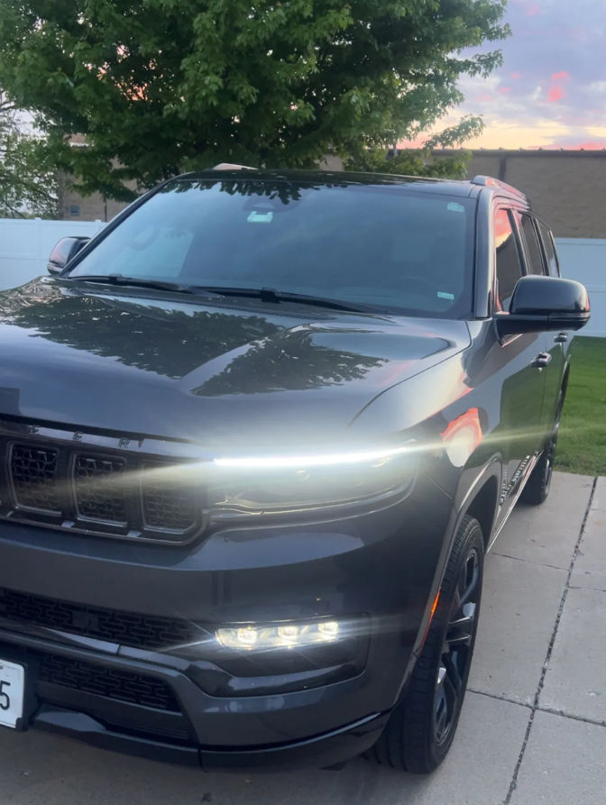 Front three-quarter view of a dark gray SUV parked on a suburban driveway at dusk, LED daytime running lights glowing with tree and white fence reflected on the hood.