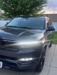 Front three-quarter view of a dark gray SUV parked on a suburban driveway at dusk, LED daytime running lights glowing with tree and white fence reflected on the hood.