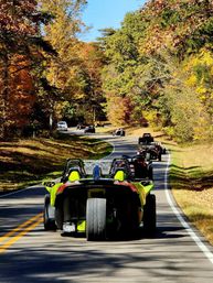 Convoy of bright three-wheeled roadsters cruising a winding country road lined with vibrant autumn foliage and clear blue sky
