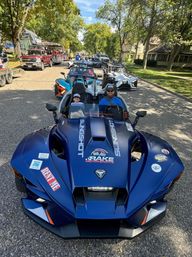 Blue three-wheeled open-air roadster with smiling adult driver and child passenger leading a lineup of sporty three-wheelers down a tree-lined small-town street during a parade