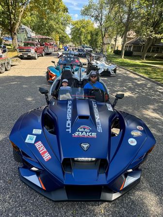 Blue three-wheeled open-air roadster with smiling adult driver and child passenger leading a lineup of sporty three-wheelers down a tree-lined small-town street during a parade
