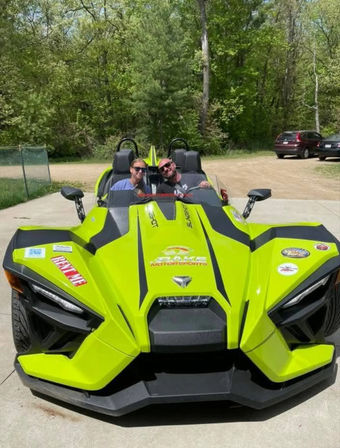 Lime-green open-top three-wheeled sport vehicle with two passengers wearing sunglasses, parked on a driveway with wooded greenery and cars in the background on a sunny day.