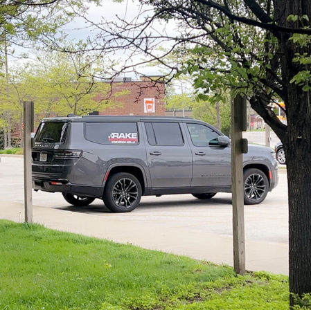 Gray full-size SUV with a company decal parked in a suburban storefront parking lot, framed by spring trees and green grass