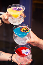 Three vibrant craft cocktails — yellow, blue, and red — served in round glasses over ice, each topped with an edible pansy flower and held by hands for a celebratory toast.