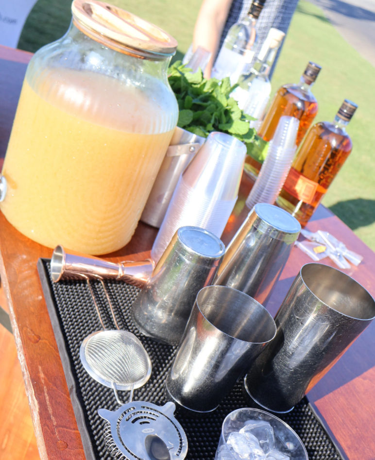 Sunny outdoor cocktail station with a large glass dispenser of orange punch, metal shakers and strainers, fresh mint, stacked plastic cups, ice and amber liquor bottles on a wooden table.