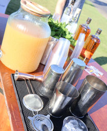 Sunny outdoor cocktail station with a large glass dispenser of orange punch, metal shakers and strainers, fresh mint, stacked plastic cups, ice and amber liquor bottles on a wooden table.