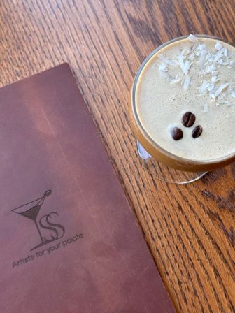Top-down shot of a frothy espresso cocktail topped with three coffee beans and coconut flakes in a coupe glass on a wood café table beside a brown menu.