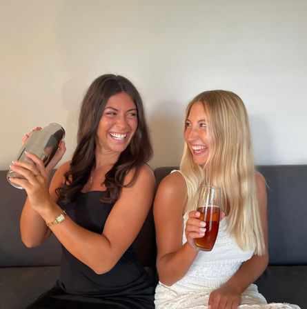 Two laughing friends on a couch in a casual indoor setting — one shaking a cocktail shaker, the other holding an amber beverage.