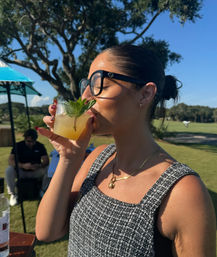 Woman in sunglasses sipping a mint-garnished citrus cocktail on a sunny outdoor lawn or golf course, wearing a sleeveless tweed top and gold necklace.
