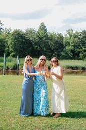 Three women in summer dresses and sunglasses toasting with wine glasses on a sunny waterfront lawn, marsh grasses and trees in the background — cheerful outdoor summer celebration.