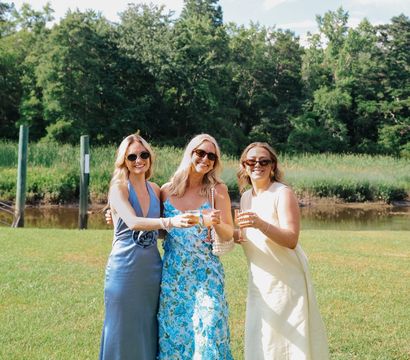 Three women in summer dresses and sunglasses toasting with wine glasses on a sunny waterfront lawn, marsh grasses and trees in the background — cheerful outdoor summer celebration.