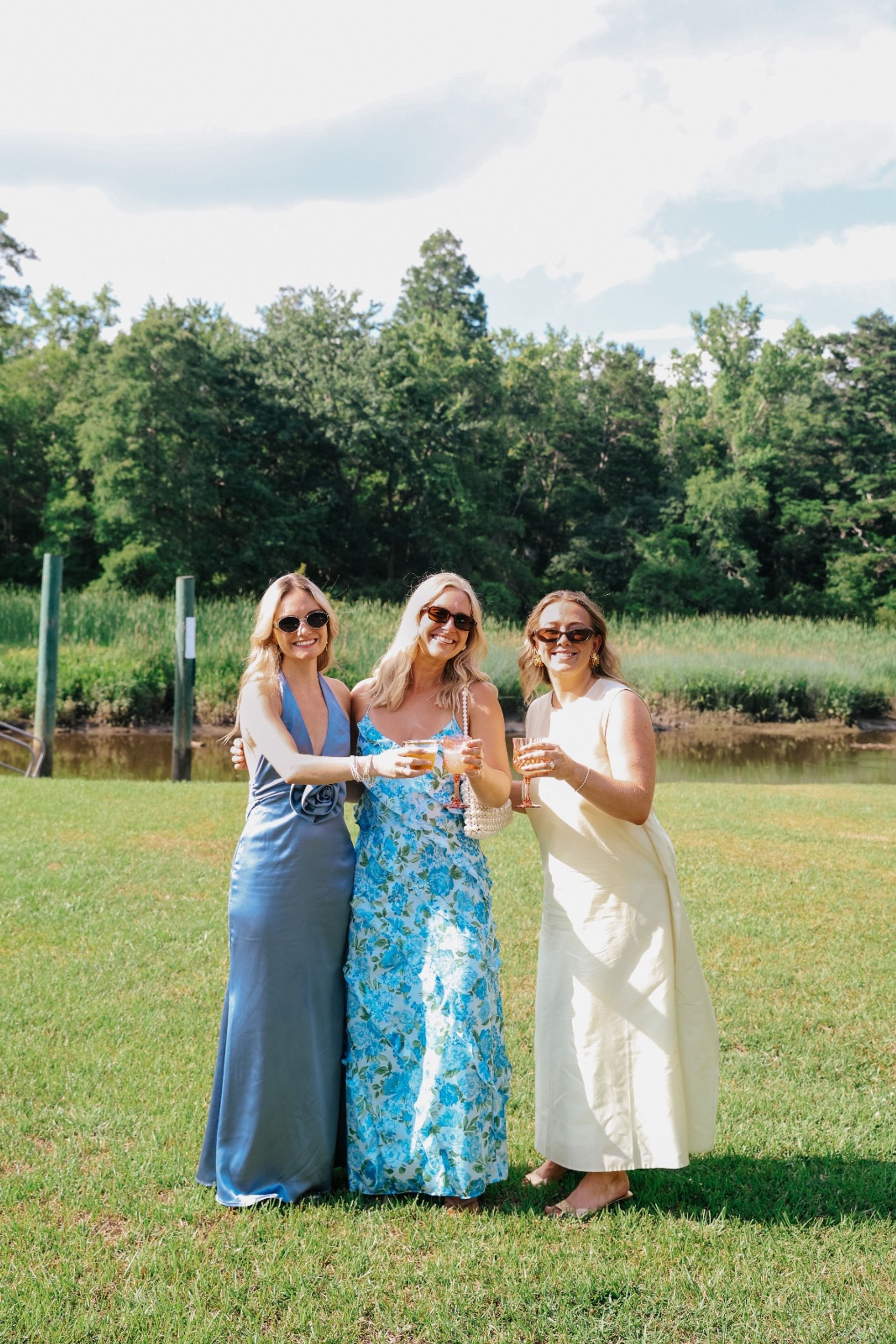 Three women in summer dresses and sunglasses toasting with wine glasses on a sunny waterfront lawn, marsh grasses and trees in the background — cheerful outdoor summer celebration.