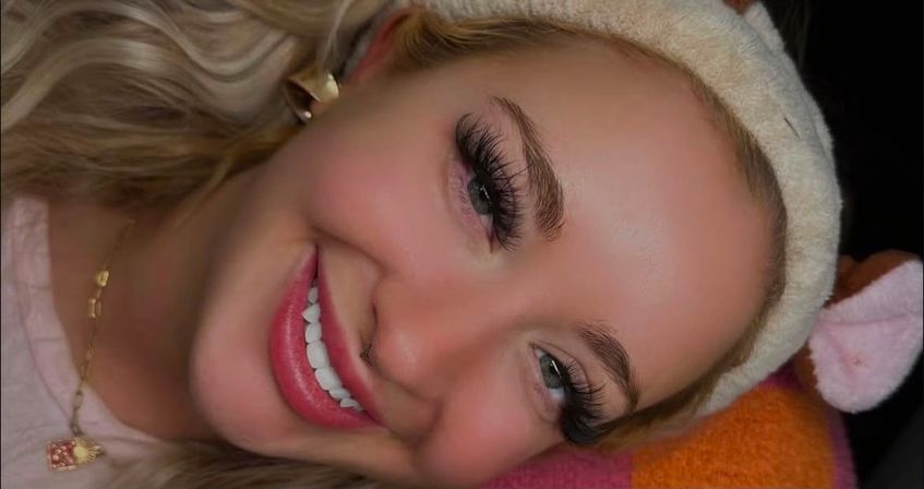 Close-up portrait of a smiling person wearing a plush headband, dramatic eyelash extensions, a gold pendant necklace, and a bright white smile — beauty lash-extension close-up.