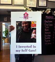Smiling client poses behind a white selfie-frame reading "I invested in my Self Care!" inside a spa/waxing salon, holding a small gift bag in front of a massage menu chalkboard and framed certificates.