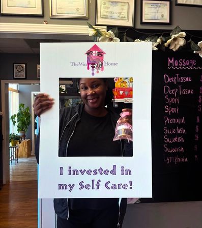 Smiling client poses behind a white selfie-frame reading "I invested in my Self Care!" inside a spa/waxing salon, holding a small gift bag in front of a massage menu chalkboard and framed certificates.