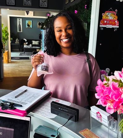 Smiling woman at a boutique front desk holding a small jar in a pink mesh gift bag, clipboard and business cards on the counter, bright pink flowers and cozy shop seating with wall art in the background.