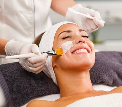 Smiling woman enjoying a spa facial skincare treatment as an esthetician brushes on a white mask