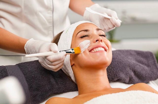 Smiling woman enjoying a spa facial skincare treatment as an esthetician brushes on a white mask