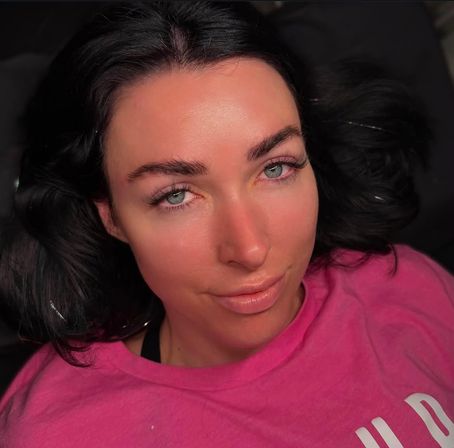 Close-up portrait of a woman lying back with dark hair, blue-green eyes, groomed brows and long lashes, wearing a pink shirt in soft indoor lighting.