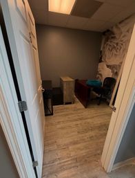 Small private office viewed through an open white door — gray walls, wood-look laminate floor, a desk with rolling chair and teal cushion, and a large grayscale rose mural.