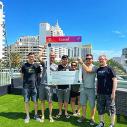 Six smiling adults on a sunny rooftop lawn in South Beach, Miami holding a large Instagram-style “Escaped” photo frame with palm trees and beachfront hotels in the background