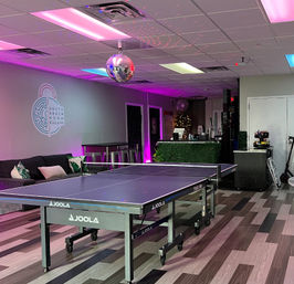 Indoor entertainment lounge with a ping pong table beneath a disco ball, neon pink and purple accent lighting, sofa seating, service counter and patterned wood-look flooring.