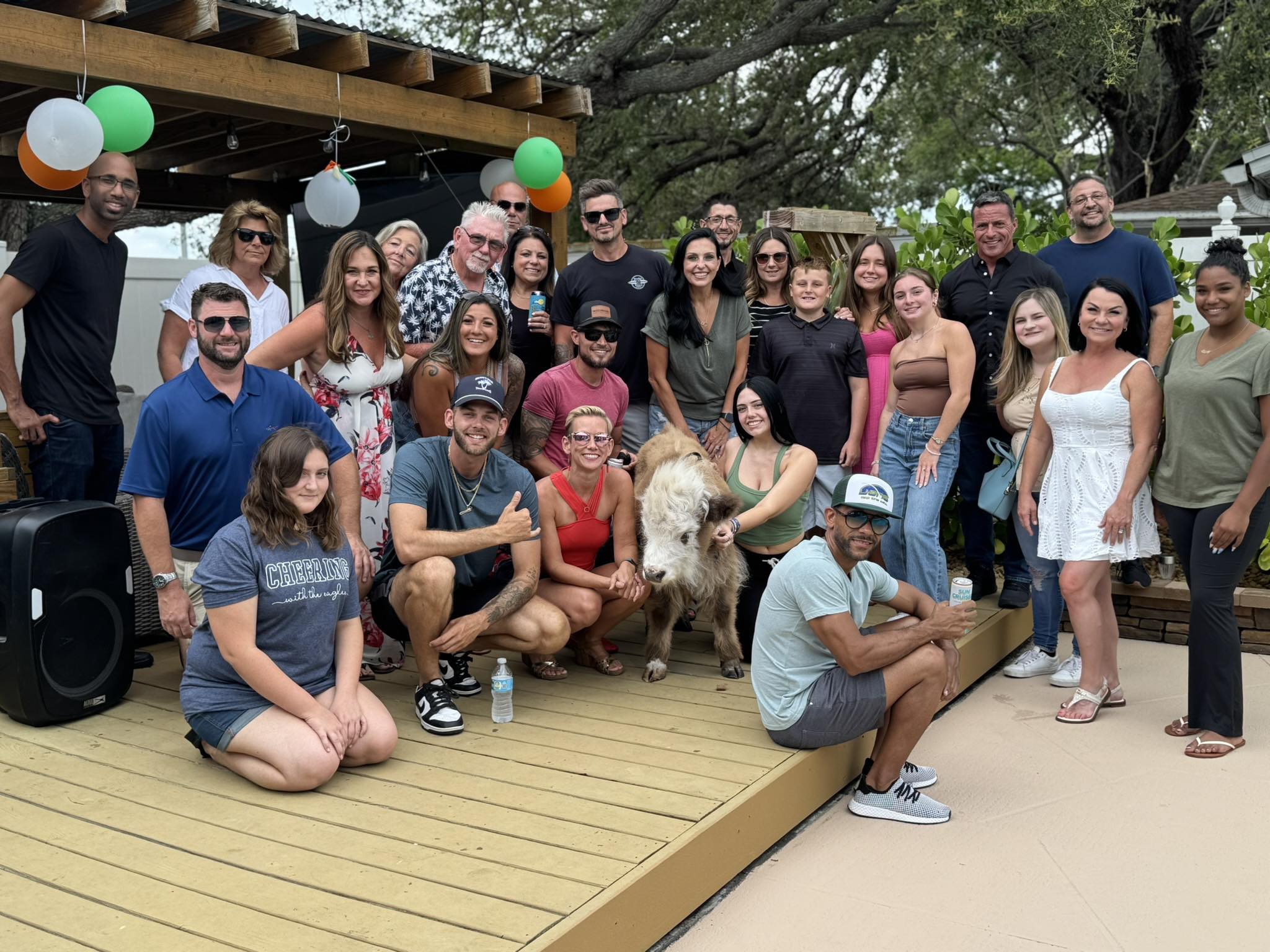 Cheerful group photo on a backyard deck—about two dozen adults and teens smiling around a small brown-and-white calf under a wooden pergola decorated with balloons.