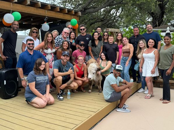 Cheerful group photo on a backyard deck—about two dozen adults and teens smiling around a small brown-and-white calf under a wooden pergola decorated with balloons.
