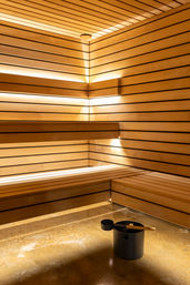 Warm, modern wood-paneled indoor sauna with tiered benches, recessed LED strip lighting and a black water bucket with ladle on the polished floor — spa wellness room.