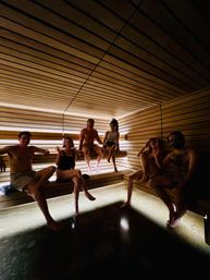 Six adults relaxing in a wood-paneled indoor spa sauna with warm recessed lighting, tiered benches and a modern minimalist design