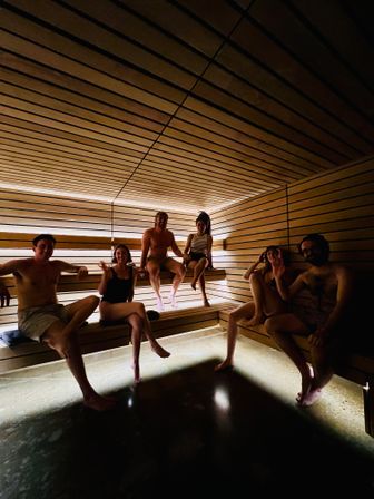 Six adults relaxing in a wood-paneled indoor spa sauna with warm recessed lighting, tiered benches and a modern minimalist design