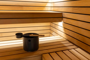 Cozy modern wooden sauna interior with horizontal slatted benches, warm ambient lighting, and a black sauna bucket and ladle on the bench