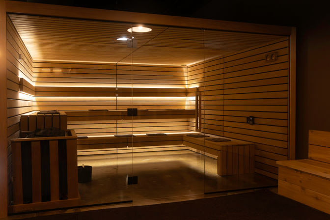 Modern indoor wooden sauna with glass door, tiered benches, heater with stones and bucket, and warm recessed LED strip lighting creating a cozy spa atmosphere.