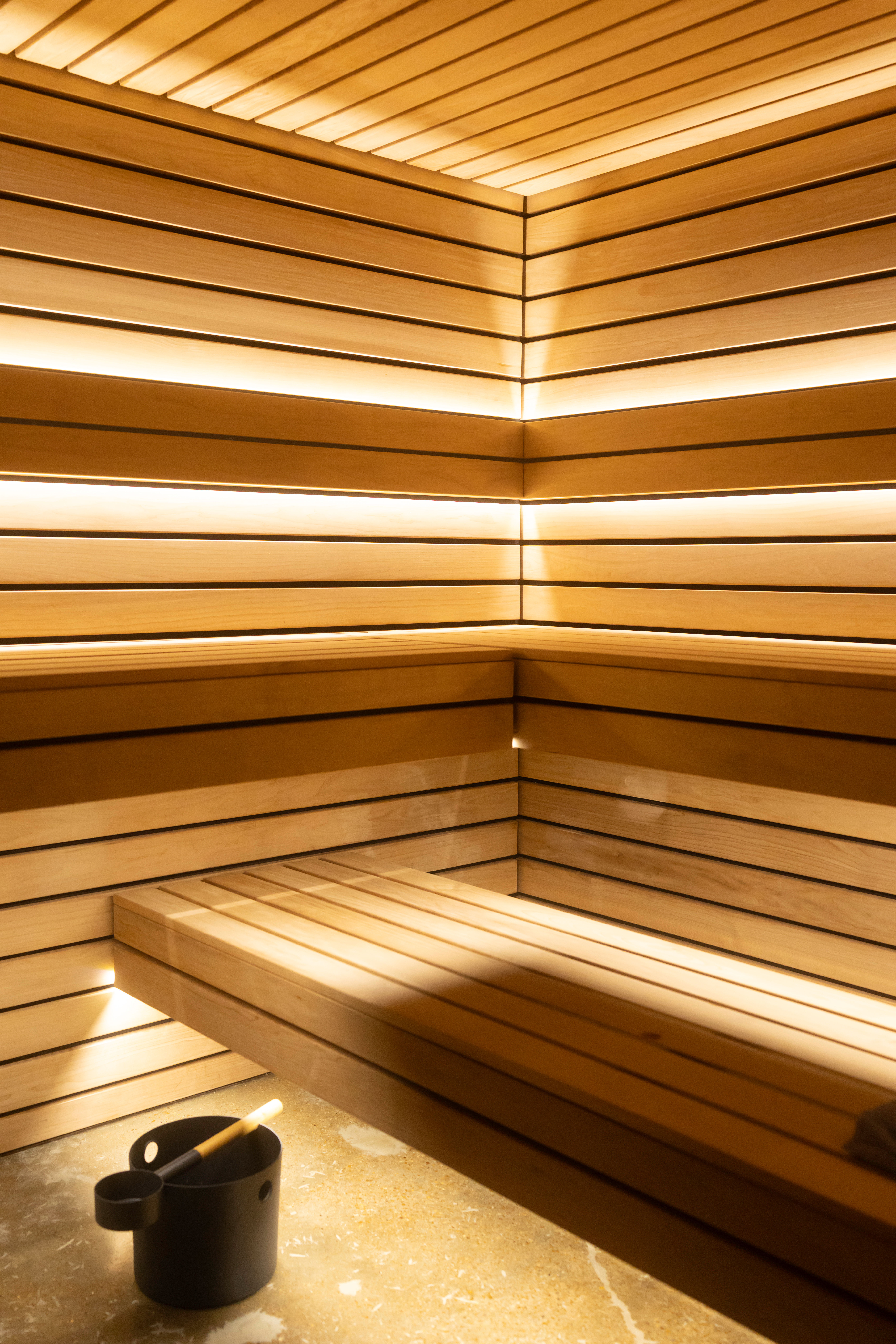 Warm modern wooden sauna interior with slatted corner benches, soft LED backlighting and a black water bucket with ladle on the floor — cozy spa wellness setting