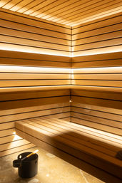 Warm modern wooden sauna interior with slatted corner benches, soft LED backlighting and a black water bucket with ladle on the floor — cozy spa wellness setting