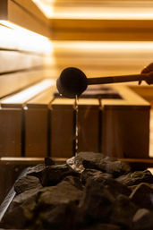 Ladle pouring water onto hot sauna stones in a wooden sauna, steam rising under warm amber lighting