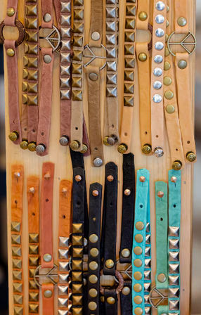 Handmade studded leather bracelets in tan, peach, black and turquoise arranged in vertical rows on a wooden display with metal studs and round buckles — boho accessory market style
