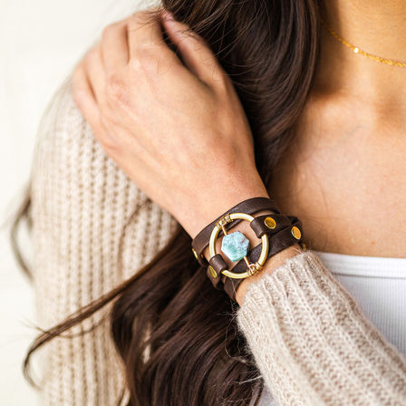 Close-up of a wrist wearing a brown leather wrap bracelet with a brass ring and aqua-blue gemstone, styled with a cozy knit sweater and long dark hair — boho fashion accessory.