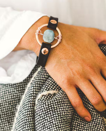 Close-up of a hand wearing a stylish black leather bracelet with a rose-gold circular frame and raw aquamarine stone, resting on a textured black-and-white woven blanket.