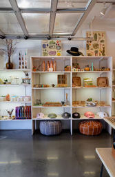 Bright boutique home-goods shop interior with white cubed shelving displaying hats, leather poufs, copper water bottles, pottery, candles, plants and botanical prints.