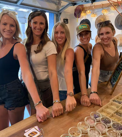 Five smiling women at a seaside boutique showing bracelets over a wooden counter with bowls of crystals and jewelry on display