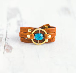 Brown leather wrap bracelet with a bright turquoise bead set in a gold circular accent and gold studs, boho jewelry on white distressed wood background