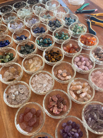 Close-up of colorful polished gemstones and crystal chips sorted in clear glass bowls on a wooden craft table, with jewelry-making pliers visible — vibrant beading and crystal supplies.