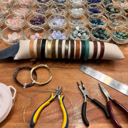 Jewelry-making workstation with rows of glass bowls holding colorful gemstone chips, leather bracelet samples on a cloth roll, coils of wire, ruler and pliers on a wooden craft table